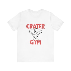 Op de afbeelding: Wit T-shirt met een rode afbeelding van een gespierde man die poseert met de tekst "CRATER GYM" in rood.