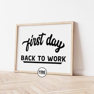Könnte beinhalten: Gerahmter Druck mit den Worten "first day" in kursiver Schrift über "BACK TO WORK" in fetter, serifenloser Schrift. Der Text ist schwarz auf weißem Hintergrund, in einem hellen Holzrahmen. Unten befindet sich ein kleines rundes Logo.
