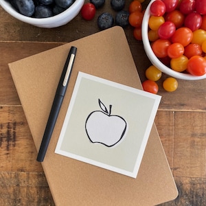 May include: A black pen rests on a brown notebook next to a small card with a black apple outline. Bowls of blueberries and red and yellow cherry tomatoes are in the background. The items are on a wooden surface.