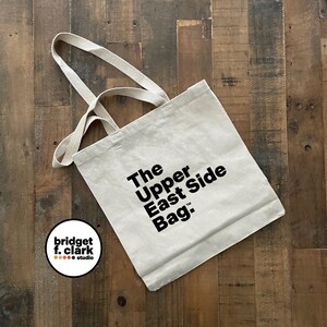May include: A white canvas tote bag with black text that reads "The Upper East Side Bag." The bag has two handles and a small logo on the bottom left corner that reads "bridget f. clark studio".