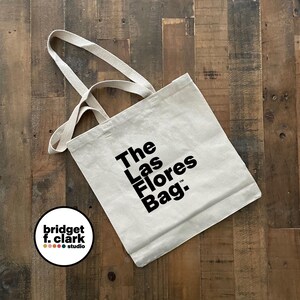 May include: Beige tote bag with the text "The Las Flores Bag." in black. The bag has long, beige handles and is made of a canvas-like material. The bag is on a wooden surface.