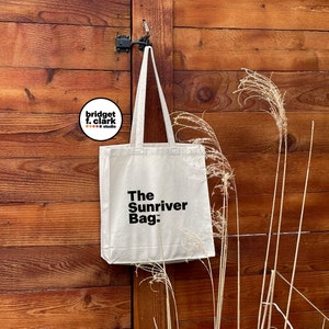 May include: A white canvas tote bag with black text that reads "The Sunriver Bag." The bag is hanging on a wooden door with a brown and black door handle.