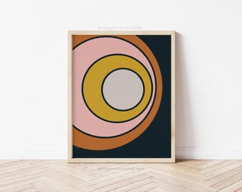 May include: A framed art print with a retro design. The artwork displays concentric circles in pink, mustard yellow, and brown against a dark blue backdrop. The print is in a light wood frame. The text "bridgett clark studio" is at the top.