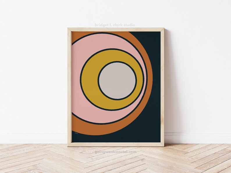 May include: A framed art print featuring a retro-style design. The artwork showcases concentric circles in shades of pink, mustard yellow, and brown against a dark blue background. The print is in a light wood frame. The text "bridgett clark studio" is at the top.