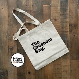 May include: A white canvas tote bag with black text that reads "The Gresham Bag." The bag has two black straps.