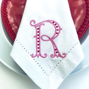 May include: A white cloth napkin featuring an embroidered pink letter R with red dot accents. The napkin has a decorative hemstitch border and is folded into a triangle shape, resting on a red plate with a beaded edge and a white plate.
