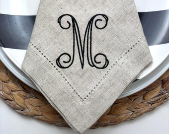 May include: A beige linen napkin featuring a decorative hemstitch border and a black embroidered monogram of the letter M in a cursive script style. The fabric has a visible woven texture and is folded into a triangular shape.