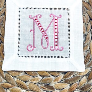 May include: A square white linen napkin featuring a central embroidered letter M in pink thread with small red accents. The napkin has a decorative hemstitched border framing the monogram, creating a clean and structured appearance for the fabric.