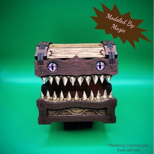 May include: A wooden treasure chest with a monster-like design. The chest is brown with sharp, white teeth and purple eyes. The text "Modeled By: Mazie" is on a brown starburst. The text "Modeling Coloring only Eyes will vary" is at the bottom.
