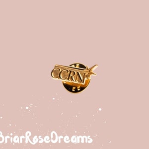 May include: Gold-colored pin with the letters "CCRN" in a rectangular shape. The pin has a small star-shaped design on the right side.