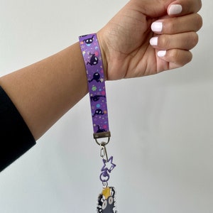 May include: Purple and white lanyard with a black and white cartoon character charm. The charm is attached to a silver star charm and a silver chain.