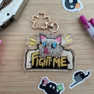 May include: A clear acrylic keychain featuring a cartoon character with a boar-like snout and the text "FIGHT ME". The character has pink cheeks, blue eyes, and gray fur. The keychain is attached to a gold metal ring.