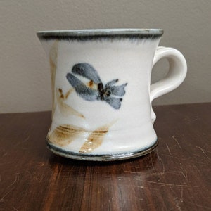 May include: White ceramic mug with a floral design in blue and tan. The mug has a curved handle and a dark rim. The floral design is hand-painted, giving the mug a unique, artistic look. The mug is sitting on a dark wooden surface.