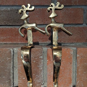 May include: A pair of gold metal reindeer stocking holders. The reindeer are stylised and have a whimsical design. They are mounted on a brick wall.