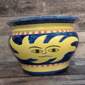 May include: A hand-painted ceramic planter with a whimsical sun face design. The planter features a yellow sun with a smiling face against a blue background. Red dots encircle the top and bottom of the yellow section. The rim is blue.