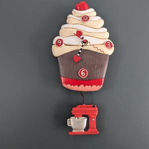 May include: A novelty cupcake-shaped wall clock with a red cherry. The clock has a brown base, cream-coloured frosting, and a red and black heart-shaped clock hand. A red mixer hangs below the clock.