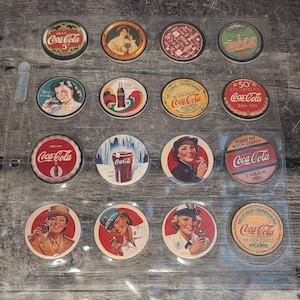 May include: A selection of vintage Coca-Cola bottle caps displayed in a clear plastic sheet. The caps showcase diverse designs, including the Coca-Cola logo, vintage adverts, and images of women. The caps are circular and feature a variety of colours.