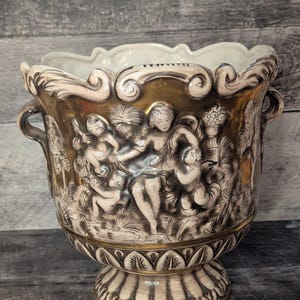 May include: A white ceramic planter with gold accents and a raised design of three figures. The planter has two handles and a pedestal base.
