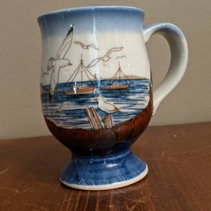 Peut inclure: Une tasse en céramique bleue et blanche avec une scène de bord de mer représentant des mouettes et des bateaux. La tasse a une bordure brune et une base bleue.