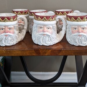 May include: Set of six white ceramic mugs featuring a Santa Claus face and a red and gold crown. The mugs have a unique handle shaped like a Santa Claus arm.