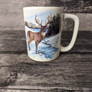 May include: White ceramic mug featuring a brown deer with large antlers standing in a snowy forest scene.
