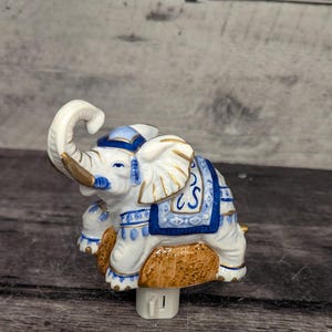 Peut inclure: Veilleuse éléphant en céramique blanche, bleue et dorée. L'éléphant a une trompe levée et est décoré d'accents bleus et dorés. La veilleuse est conçue pour être branchée sur une prise électrique standard.