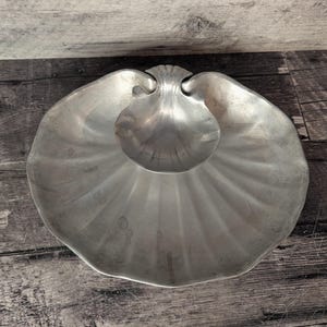 May include: A silver-coloured, shell-shaped serving dish with a smaller, shell-shaped bowl in the centre. The dish has a fluted design radiating from the centre. The dish is made of metal and sits on a wooden surface.
