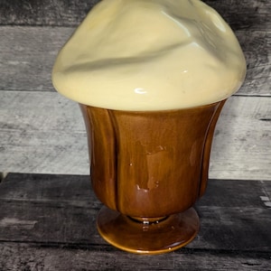 May include: A ceramic ice cream sundae dish with a white lid and a red cherry on top. The dish is brown and has a fluted design.