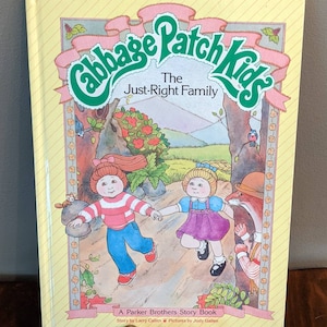 May include: A yellow book with a green and pink Cabbage Patch Kids logo. The title is "The Just-Right Family". The cover features an illustration of two Cabbage Patch Kids dolls walking in a woodland setting. The book is a Parker Brothers Story Book with a story by Larry Callen and pictures by Judy Gallen.