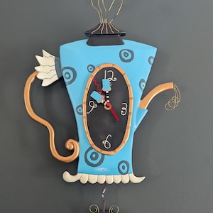 May include: A quirky wall clock in the shape of a blue teapot, adorned with black polka dots and a black clock face. It features gold accents, a white frilly edge, and a matching teacup below. The clock hands are red.