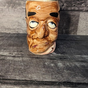 Peut inclure: Une tasse en céramique en forme de visage d'homme avec un glaçage brun et blanc. La tasse a une large ouverture ronde et une petite poignée sur le côté. Le visage de l'homme a une expression surprise avec des yeux grands ouverts et une bouche légèrement ouverte.