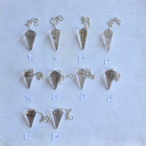 May include: Ten clear quartz crystal pendulums with silver chains hanging from a white background. The pendulums are numbered 1 through 10.