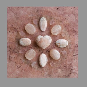 May include: A collection of polished selenite crystals arranged in a circle on a pink marble surface. The crystals are in various shapes, including oval and heart.