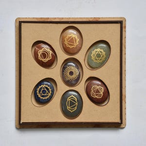 Op de afbeelding: Een set van zeven ovale stenen in een houten bakje. Elke steen heeft een andere kleur, waaronder bruin, blauw en groen, en heeft een gouden geometrisch ontwerp. De stenen zijn in een cirkelvormig patroon in het bakje gerangschikt.