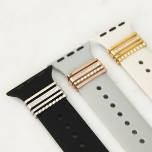 May include: Close-up of three Apple Watch bands in black, gray, and white. Each band features a decorative metal accent with a row of clear rhinestones. The metal accents are silver, rose gold, and gold.