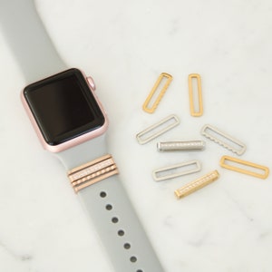 May include: A rose gold Apple Watch with a gray band, accessorized with decorative bands and gold and silver strap keepers. Several gold and silver strap keepers are scattered around the watch, some with crystal accents.