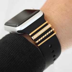 May include: A black smartwatch band with gold-colored decorative accents. The gold accents include three horizontal bars, one of which is adorned with small, clear crystals. The watch face is silver.