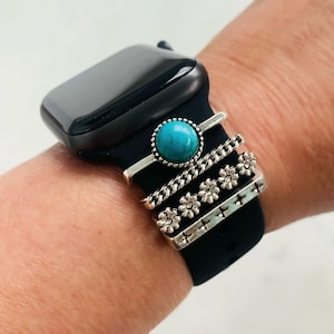 May include: Black smartwatch with a black silicone band. The band is adorned with silver-toned decorative rings. One ring features a turquoise stone, while others have chain, floral, and cross designs. The watch is on a wrist.