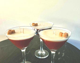 May include: Three martini glasses filled with a dark reddish-brown liquid topped with a thick layer of cream-coloured wax. Each glass features a central wick and small, light brown, cube-shaped wax embeds resembling toppings on the surface.