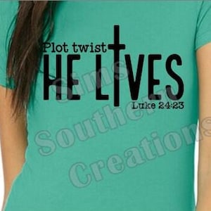 May include: Teal t-shirt with the text "Plot twist: HE LIVES" in black, with a cross incorporated into the "L". The text "Luke 24:23" is below. The shirt is a crew neck.