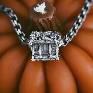 May include: A silver charm shaped like a small house with the word "BOOKSTORE" above the door. The charm is attached to a silver chain and is resting on top of an orange pumpkin.
