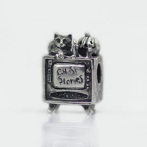 Puede incluir: Un charm de plata que representa un televisor retro con las palabras "Ghost Stories" en la pantalla. Un gato y una calabaza tallada están encima. El charm tiene un tema de Halloween.