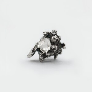 May include: A silver charm in the shape of a small, detailed, textured tree with a hollow center.