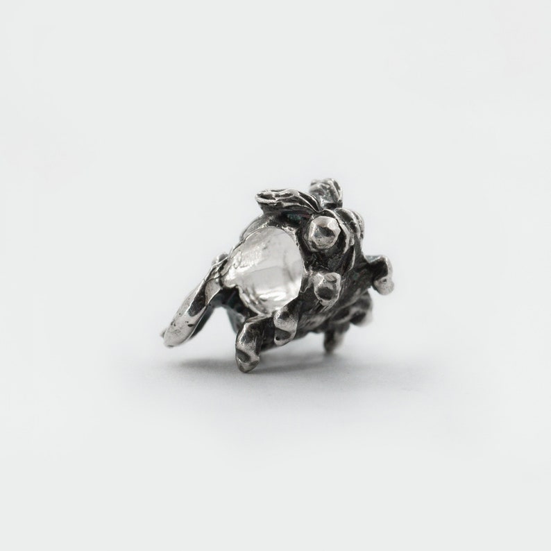 May include: A silver charm in the shape of a small, detailed, textured tree with a hollow center.