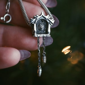 May include: A silver-toned pendant necklace featuring a miniature cuckoo clock design. The clock has intricate details, including a clock face and dangling chain with decorative weights. The necklace is on a silver chain.
