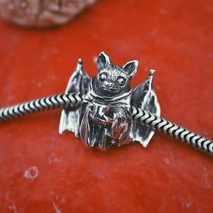 May include: A silver bat charm with detailed wings and a textured body. The bat is holding a small object, and is strung on a silver chain. The charm is set against a red background.
