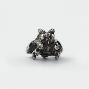 May include: A silver charm in the shape of a small, detailed creature with wings. The creature has large, round eyes and a menacing expression.