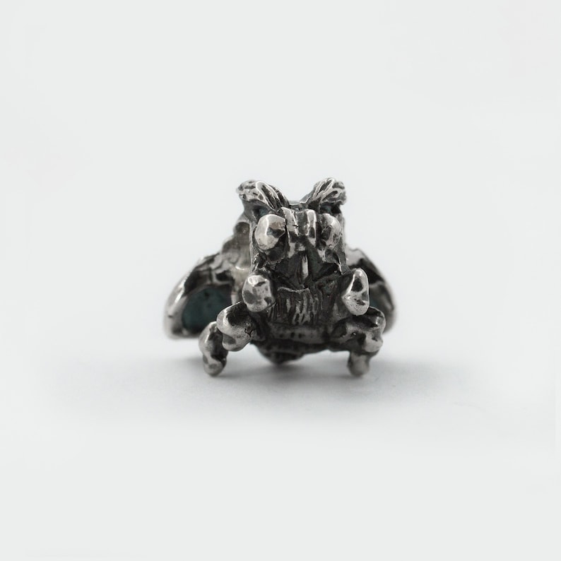 May include: A silver charm in the shape of a small, detailed creature with wings. The creature has large, round eyes and a menacing expression.