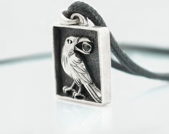 May include: A silver pendant featuring a detailed raven design with a black gemstone eye, set in a square frame. The pendant is attached to a black cord.