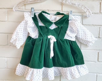 Puede incluir: Vestido de bebé vintage con corpiño de terciopelo verde y mangas y ribete de falda de lunares blancos. El vestido tiene un cuello blanco y un lazo blanco en la cintura. El vestido está colgado de una percha de plástico transparente.
